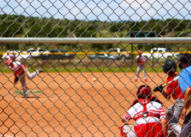 View from behind the fence  of a pitch for a baseball game with a team dressed in red and one in white at bat.