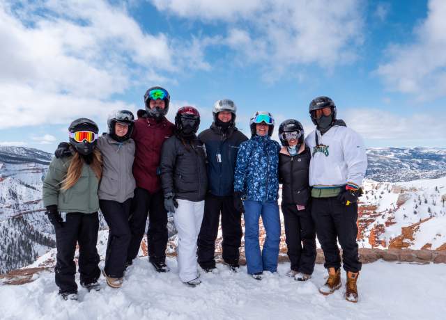 A group of eight adults dressed for winter recreation stand together for a photo at the North Overlook of Cedar Breaks National Monument