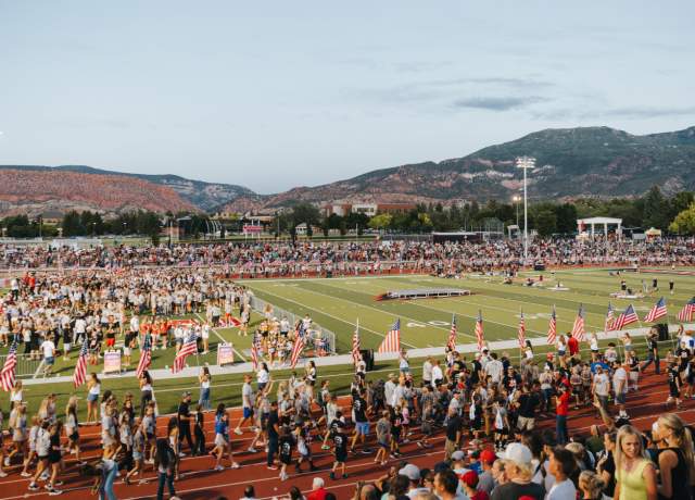Large crowd gathered at a Cedar City football stadium with American flags and red rock mountains in the background at sunset