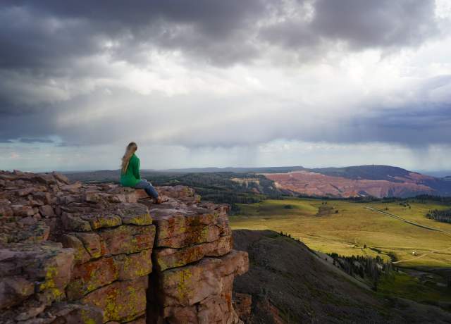 Woman in green jacket sits on the rock outcropping of the Brian Head Peak overlook at 11,000 feet elevation with Cedar Breaks National Monument in the background.