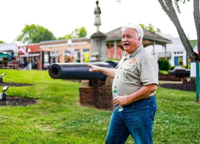 Man speaking on grass with a cannon and a building in the background