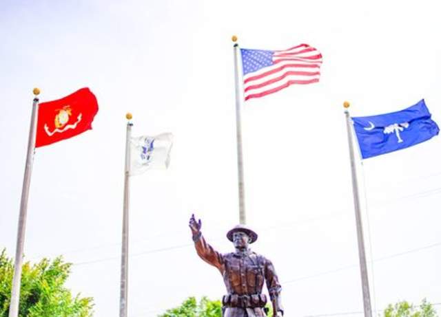 Metal statue of a military man with his arm extending, four flags including the american flag are seen in the background