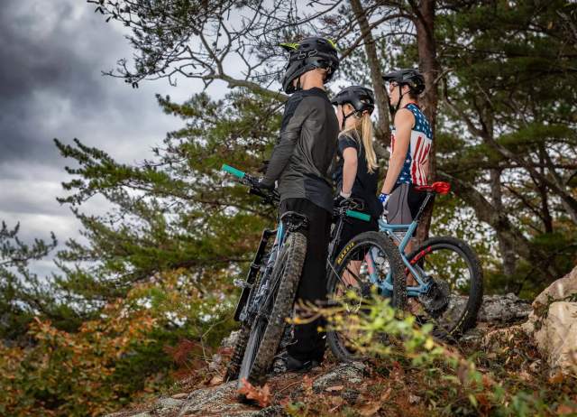 Three young mountain bikers stare out over the view of an overlook during a cloudy day at Double Oak Park. They each are standing next to their mountain bikes.