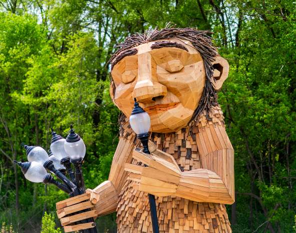 A large wooden sculpture of 'Mama Rosa,' a Thomas Dambo troll installation in Wauwatosa. The friendly giant figure is constructed from reclaimed wood pieces in natural tones, featuring a carved face with a gentle smile and twig-like hair made from bundled branches. The troll holds a bouquet of old-fashioned street lamps in one hand while lifting one lamp to her nose. The sculpture is set against a lush green forest backdrop with dense tree canopy, creating a whimsical scene that blends art with nature.