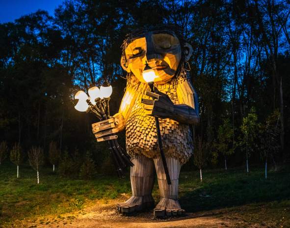The Thomas Dambo Troll, Mama Rosa is a large wooden sculpture which stands outdoors at dusk, illuminated by warm lights it holds in its hands. The figure is made of textured wooden planks, with a gentle, expressive face, and is surrounded by dark trees in the background.