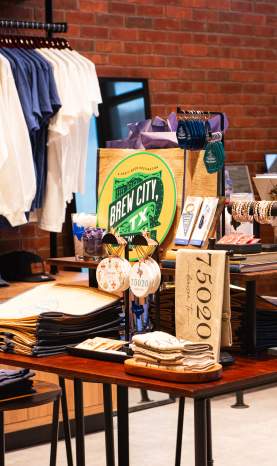 Visitor Center Interior of Store with shirts, tea towels, ornaments, bracelets, keychains, shirts, books, tote bags, pins, and Brew City Designation sign on the table.