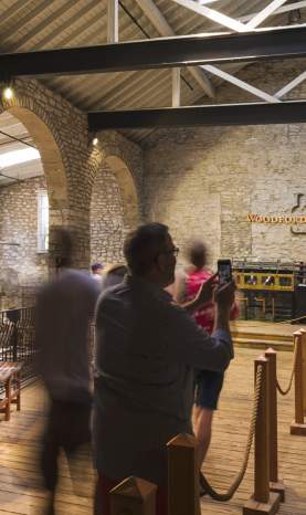 A candid photo of people taking a tour of one of the rooms of Woodford Reserve’s Distillery Tour. It’s a tall room with white brick walls, and three large bronze-colored metal devices used in the bourbon production process. The Woodford Reserve name and logo can be seen straight ahead on the wall.
