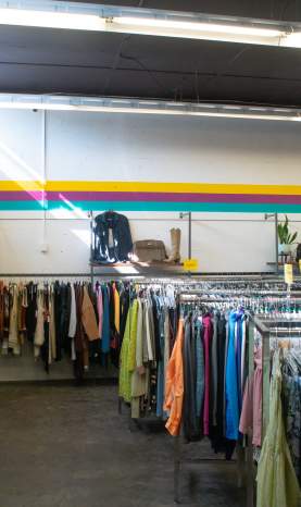 Rows of colourful secondhand clothing, including jeans, jackets, shirts, and dresses, fill a thrift store with bright overhead lighting and yellow, purple, and teal stripes along the walls.