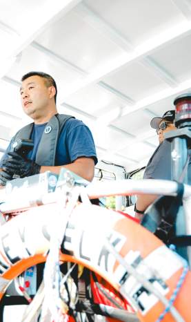 Two crew members wearing life vests operate a small ferry boat, with a life ring and safety gear in the foreground.