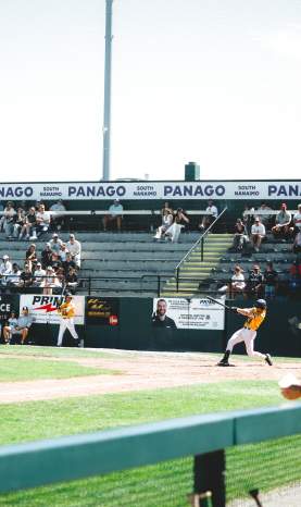 A batter swings at the pitch during a baseball game while teammates and spectators watch from the dugout and bleachers.