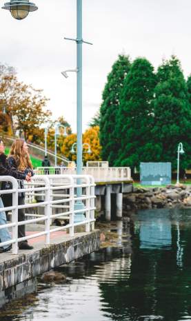 Two people pause along Nanaimo’s waterfront walkway, leaning on a railing and enjoying the view of the calm water and nearby park.