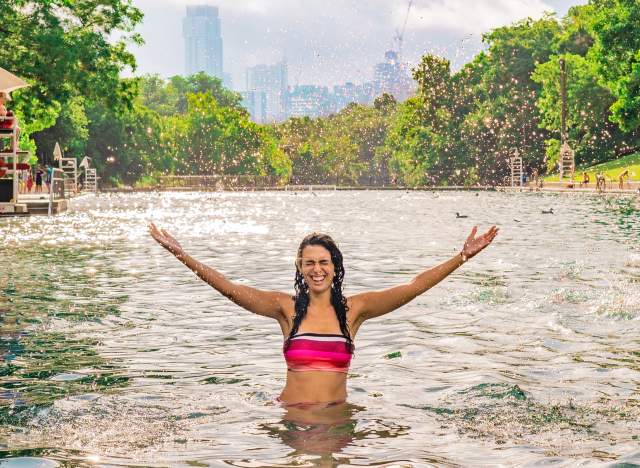 Woman standing in Barton Springs Pool wearing pink bathing suit hand holding hands up after splashing water. Austin city skyline is behind her in the distance