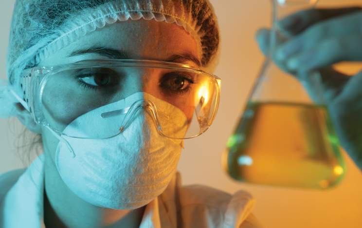 Woman wearing mask and goggles and looking at a beaker of yellow liquid.