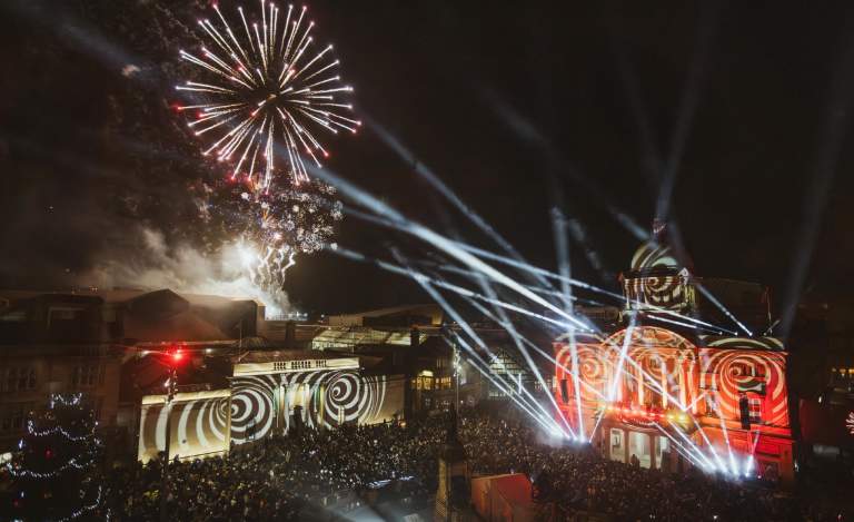 An overview of Hull City Hall and the Maritime Museum, lit up at night with spirals and flashlights, whilst fireworks explode overhead. A large crowd is gathered to watch the display.