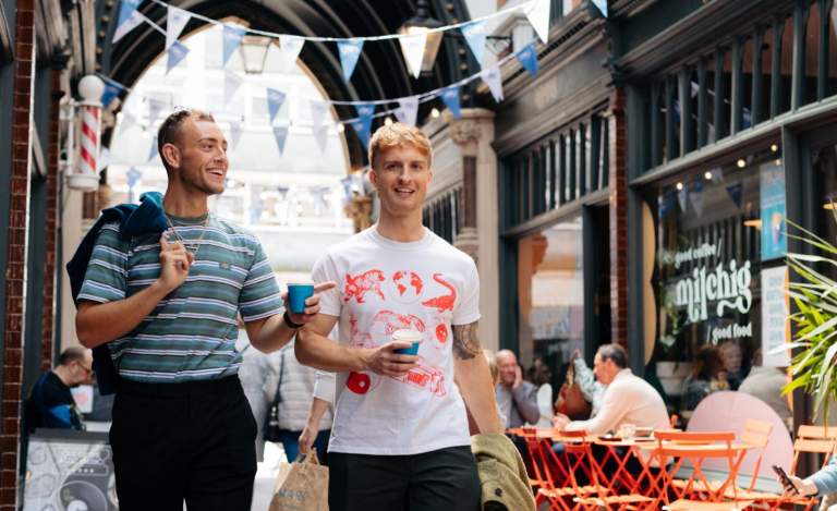 Two man strolling Paragon Arcade on a summer's day. The are holing takeaway coffees and smiling. The man on the left is wearing a blue striped t-shirt and black trousers with a dark jacket over his shoulder. The man on the right is wearing a white t-shirt with neon prints of animals on and dark trousers. Blue bunting is hung from the beamed ceilings. People are sat outside a café with orange seating and plants.