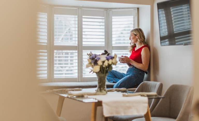 A girl in a red top sits on the inside of a set of bay windows, holding a mug and looking outside smiling. She has a table of flowers in front of her.