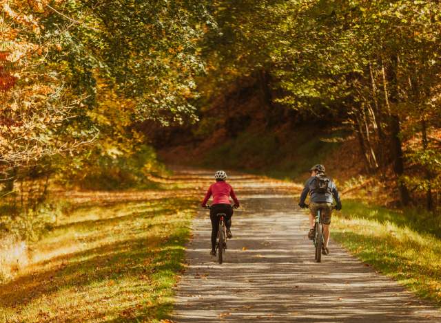 A couple ride bike on a gravel path surrounded trees with fall colored leaves of yellows, oranges, and reds.