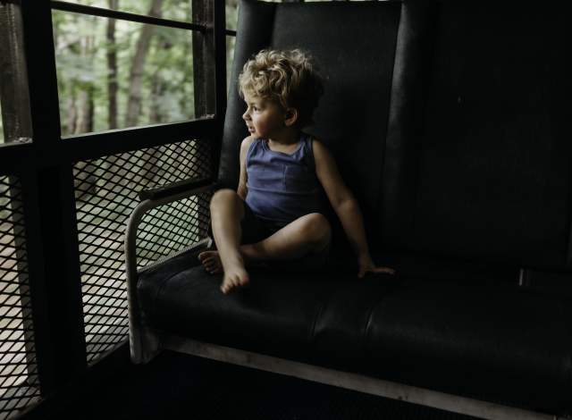 A blonde haired boy in a blue shirt and shorts stares out the window of a train.