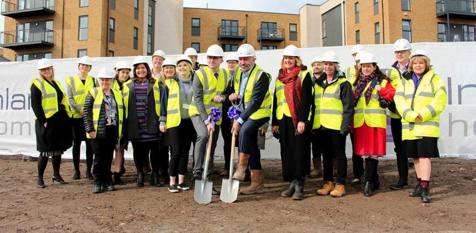 A group of people wearing hig vis and hard hats on a new development site. The two people in the middle have spades they are posing with for the camera