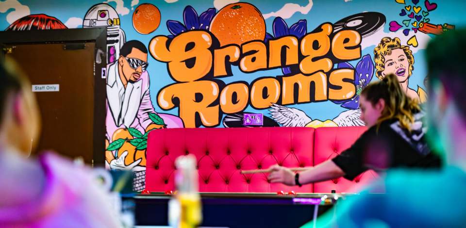Someone playing pool inside Orange Rooms with a bright pink sofa in the background and an Orange Rooms sign on the wall