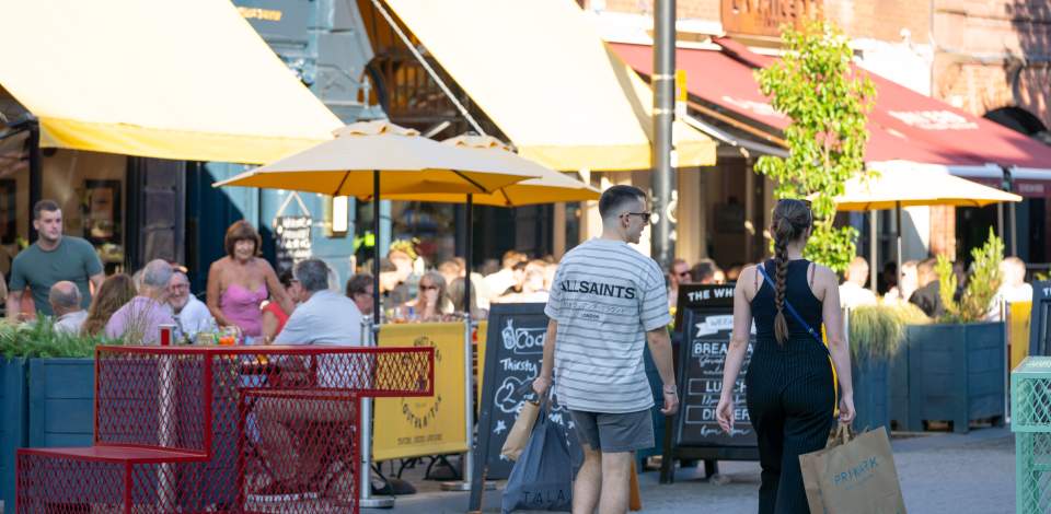 Couple walking down Oxford Street with shopping bags on a sunny day as people dine on outdoor terraces