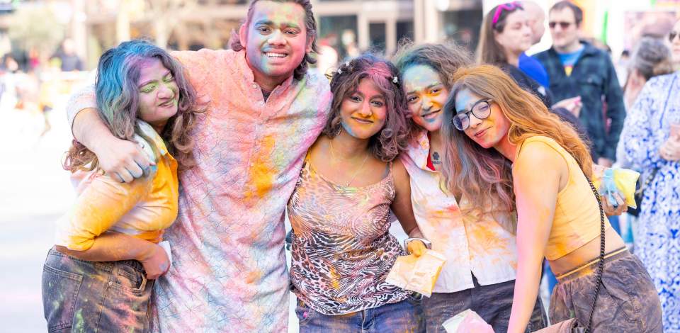 5 people with their arms around each other covered in colourful powder smiling at the Holi event in Guildhall Square