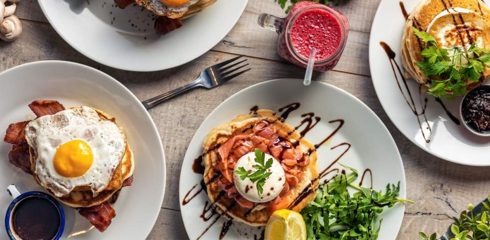 Spread of fluffy pancakes topped with breakfast toppings such as eggs and bacon, with a berry smoothie on the side
