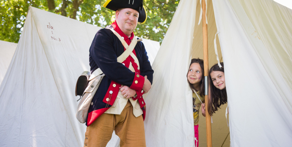 A Colonial reenactor poses in front of a tent, while two children peek out from inside