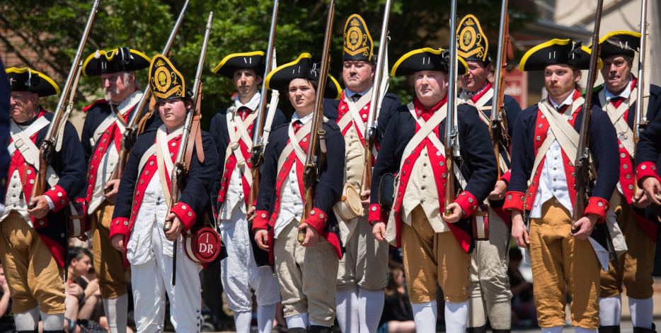 Historical reenactors in Colonial American military uniforms, holding muskets, standing in formation outdoors.