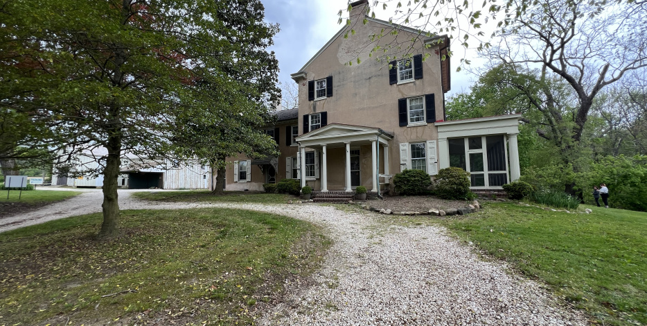 Two-story historic house with a portico entrance, set back from a gravel driveway, with a white barn visible to the left and trees around.