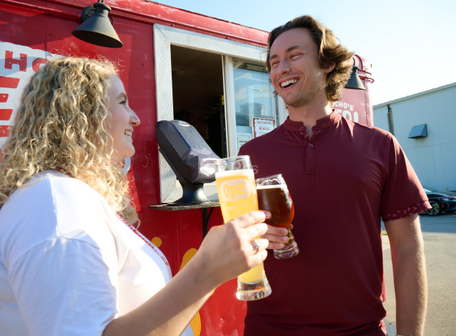 Two friends smile while holding pints of beer outside a red food truck, enjoying drinks together on a sunny afternoon at a brewery.