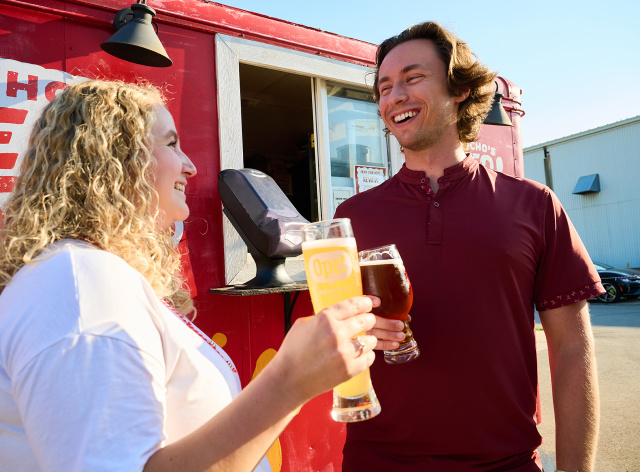 Two friends smile while holding pints of beer outside a red food truck, enjoying drinks together on a sunny afternoon at a brewery.