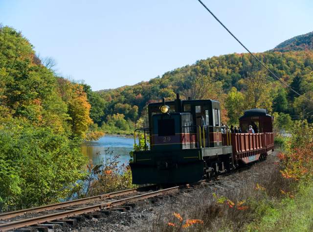 A vintage train travels along tracks beside a serene river, surrounded by lush autumn foliage under a clear blue sky. Passengers ride in open-air cars.