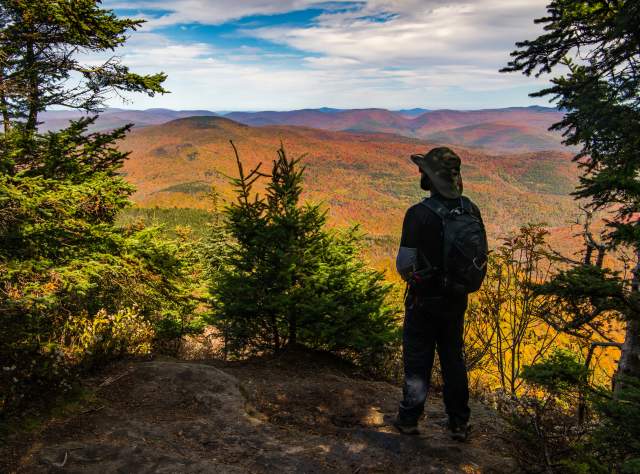 A person with a backpack stands on a rocky ledge overlooking a vast landscape of autumn foliage. Evergreen trees frame the view under a partly cloudy sky.