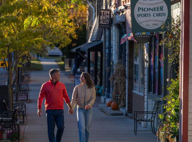 A couple walks hand-in-hand down a quaint, sunlit street lined with shops in autumn. Leaves are turning, pumpkins decorate storefronts, and they seem content.
