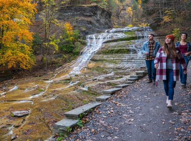 Three people hike along a scenic trail next to a tiered waterfall in autumn. The path is lined with colorful foliage and fallen leaves.