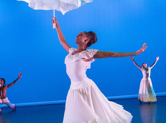 Two female and one male dancer dressed in white dance in front of a blue backdrop at the Alvin Ailey American Dance Theater