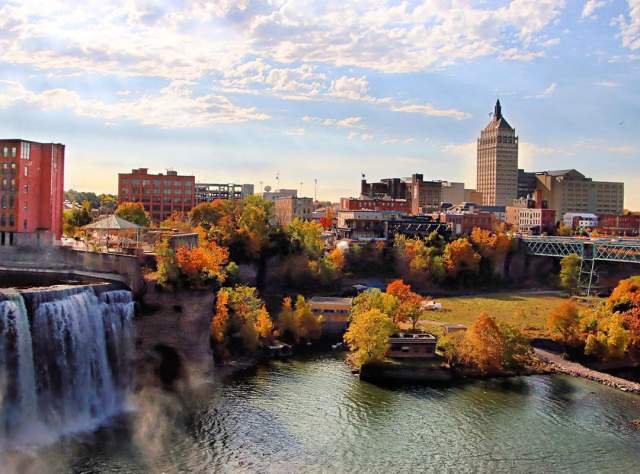 Landscape view of high falls waterfall in downtown Rochester surrounded by tall buildings, bridges, and fall foliage