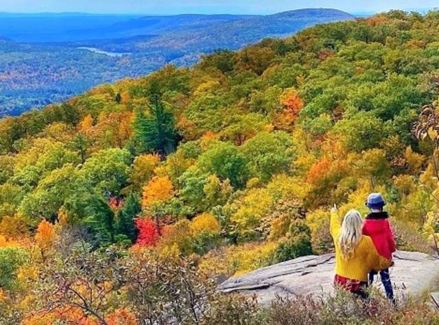 A woman points out Lake George to a child from the summit of Prospect Mountain with trees covered in various hues of fall colors below