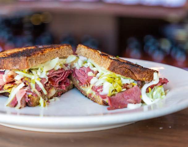 A corned beef sandwich with shredded cabbage on toasted rye, served on a white plate. The setting is cozy, with a background of blurred bottles.