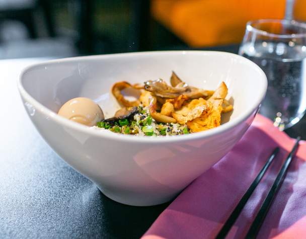 A bowl of ramen on a dark table, featuring a soft-boiled egg, mushrooms, and green onions. Nearby, there is a glass of water and black chopsticks on a pink napkin.