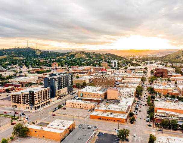 Aerial view of downtown Rapid City at sunset, with downtown streets lined by low-rise buildings, cars, and parking lots. Rolling green hills surround the city, while dramatic clouds stretch across the sky and golden sunlight glows on the horizon.