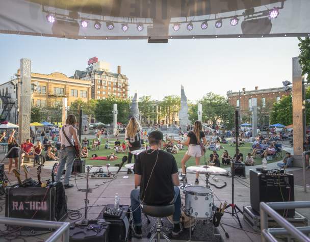 Outdoor concert scene with a band performing on stage, viewed from behind the drummer. Audience relaxes on grass, under a clear sky, enjoying the music.