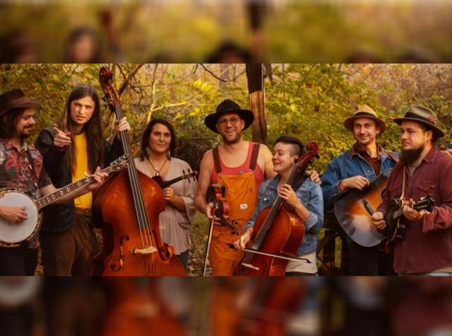 The seven musicians of The Debutants stand in a line holding their instruments in a wooded area.