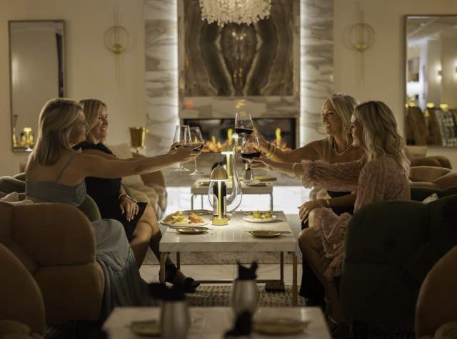 Four women raise glasses of red wine while sitting around a marble table with food in a cozy, stylish lounge.