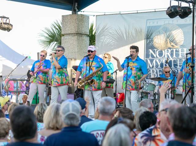 A band performs on an outdoor stage in North Myrtle Beach, wearing colorful tropical shirts and entertaining a large crowd. One musician plays a saxophone while others sing and play guitar, drums, and keyboard. The audience watches enthusiastically. A banner in the background features the North Myrtle Beach logo.