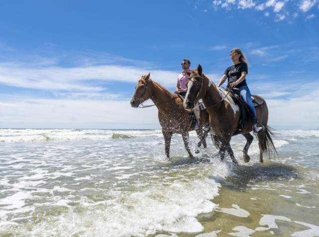 Two people horseback riding along the shoreline in North Myrtle Beach, South Carolina, with waves splashing under sunny blue skies, showcasing coastal adventures and outdoor fun.