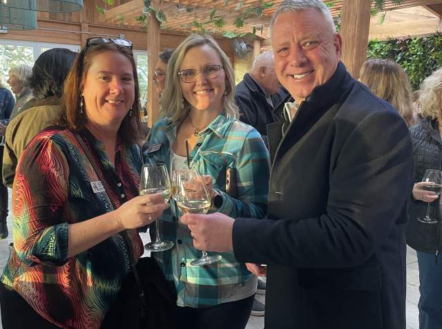 Three people smile and toast with wine glasses at an indoor event with a wooden ceiling, greenery, and hanging light fixtures. The group stands close together in a lively crowd, enjoying the social atmosphere.