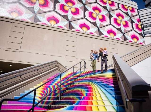 Family on stairs that are painted in a rainbow swirl with cherry blossoms on the wall behind them