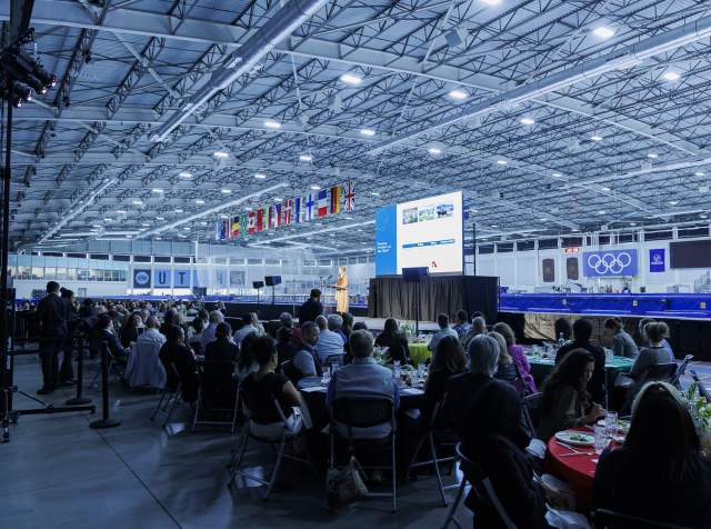 Speaker on stage at the Utah Olympic Oval, people sitting at round tables in the foreground, UT Hockey Club and Olympic logos in the background